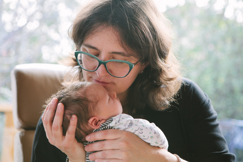 A mother is seated in front of a window, lovingly kissing her baby.