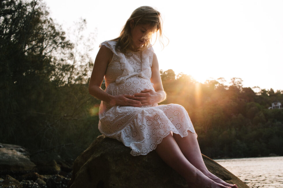 A woman who is pregnant is sitting on a rock during sunset.