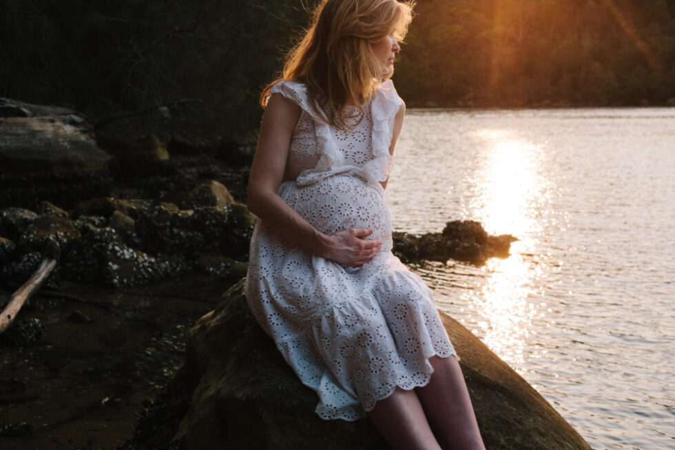 A mother-to-be sits on a rock during sunset.